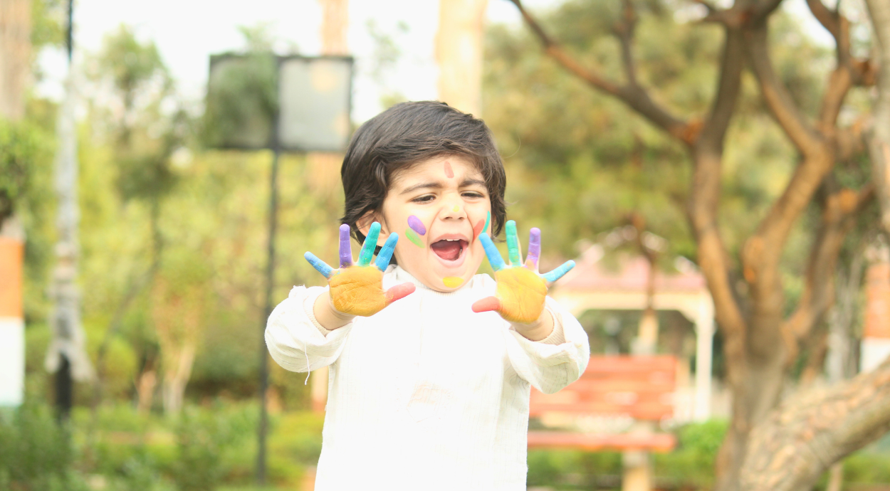 A joyful young child wearing a white outfit, playing with bright Holi colors on their hands and face in an outdoor park setting.