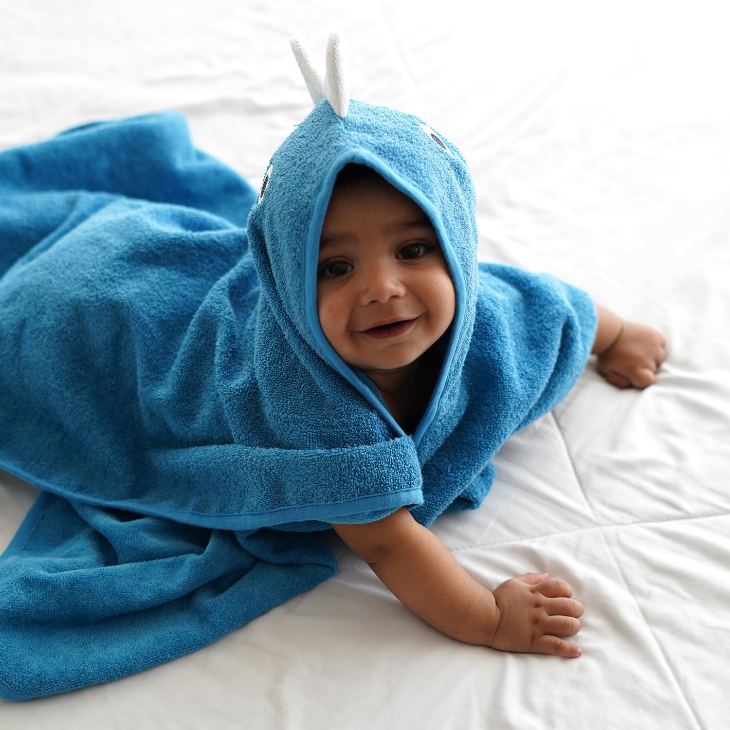 Smiling baby wrapped in a blue hooded towel with playful white horn accents, lying on a white bed. The baby is looking at the camera, with arms extended, creating a cozy and cute scene.