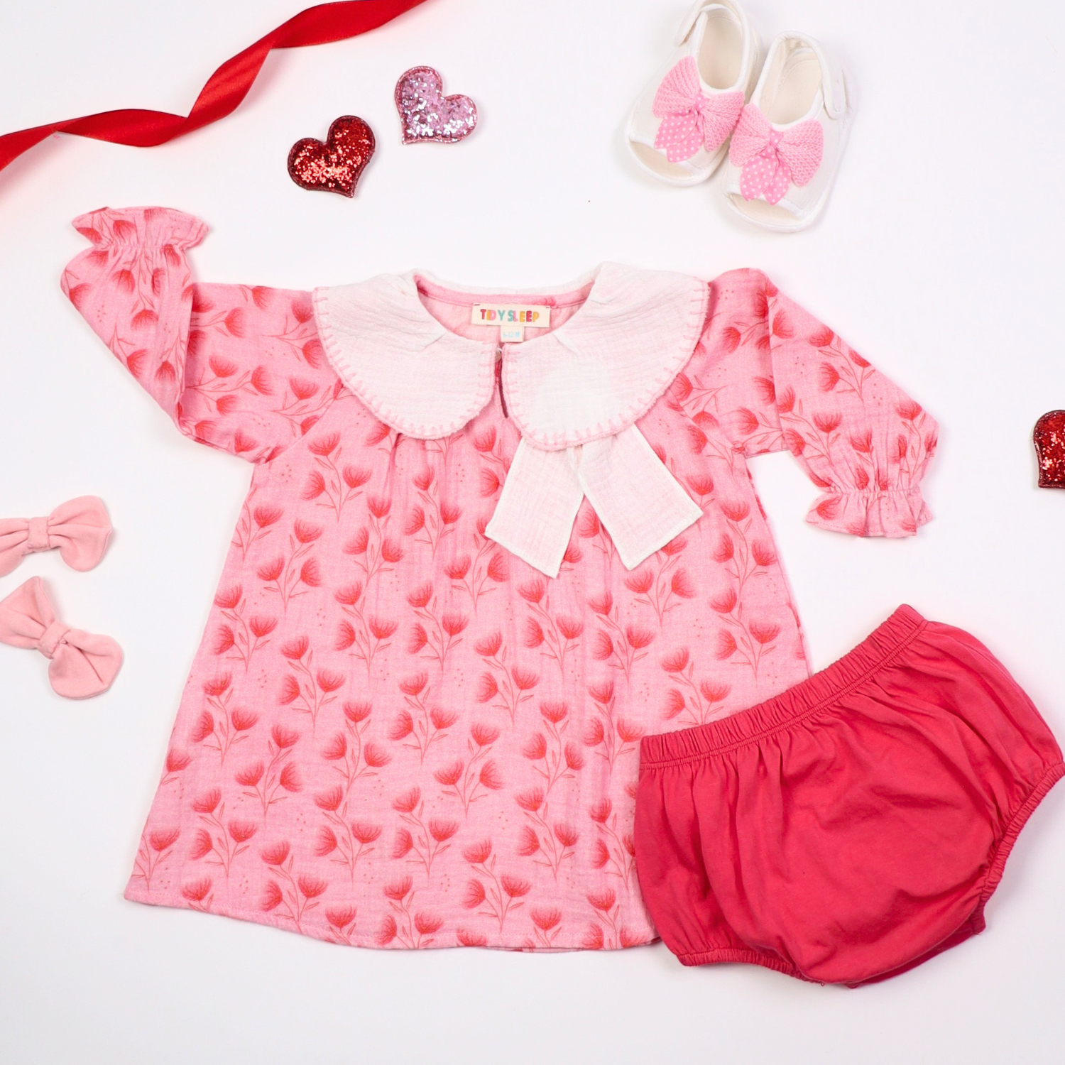 Pink floral baby dress with a large white collar and matching bloomers, displayed on a white background with decorative elements like bows, hearts, and baby shoes.