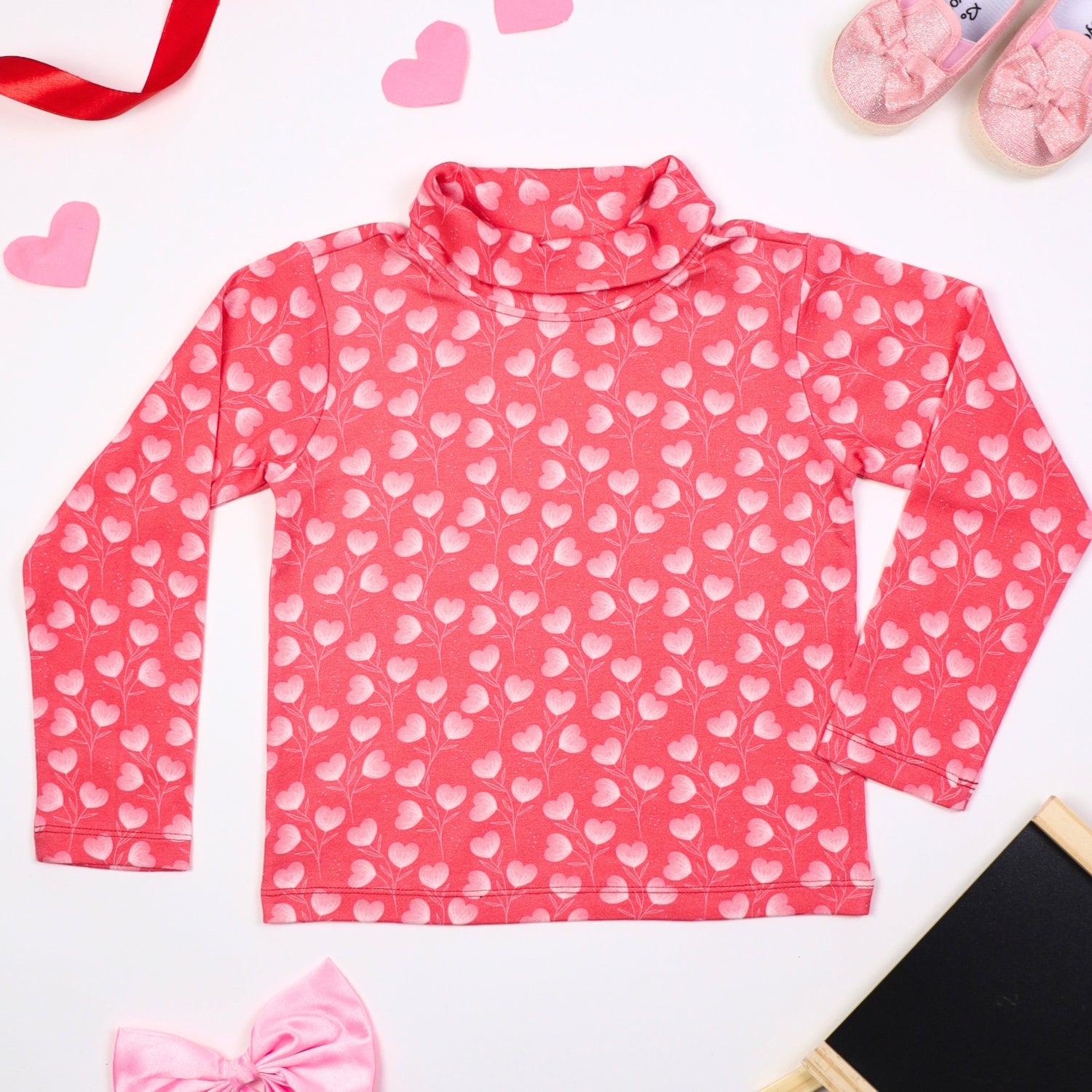 Pink turtleneck long-sleeve top with a heart print, displayed on a white background with decorative elements like ribbons, hearts, a pink bow, and baby shoes.
