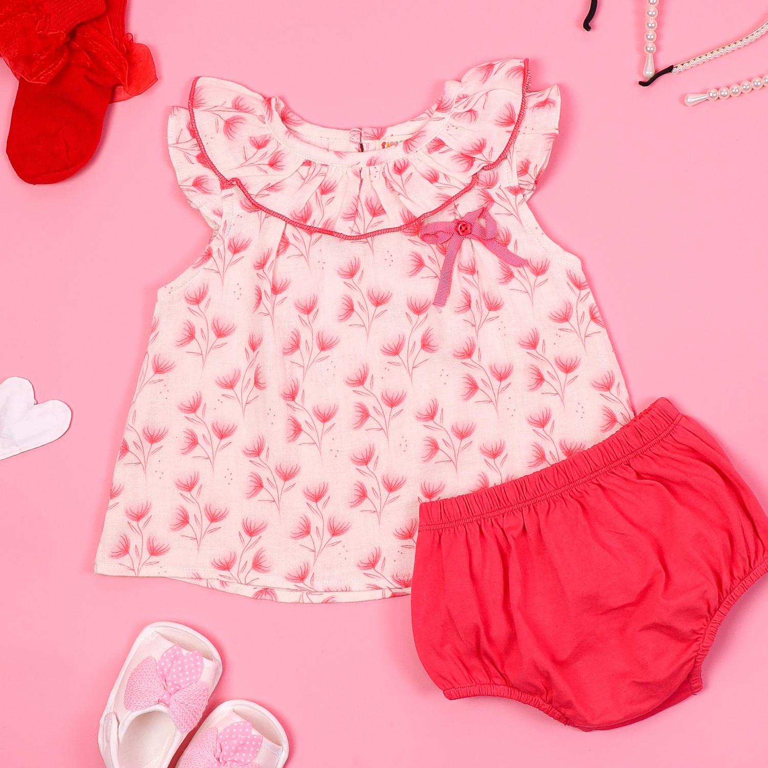 Sleeveless pink floral baby dress with a ruffled collar and matching bloomers, displayed on a pink background with accessories like baby shoes, socks, and pearl necklace.