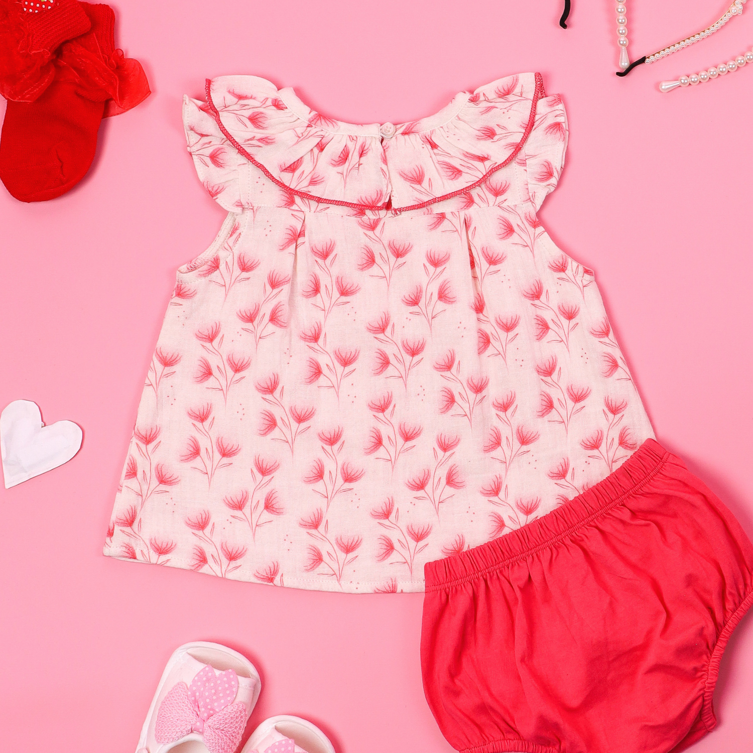 Back view of sleeveless pink floral baby dress with a ruffled collar and matching bloomers, displayed on a pink background with accessories like baby shoes, socks, and pearl necklace.