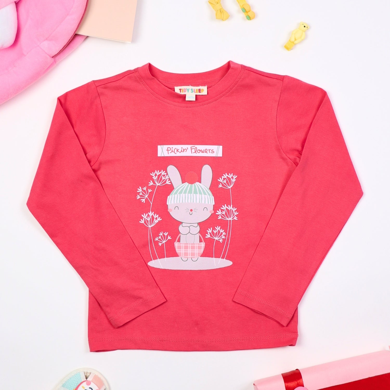 Pink long-sleeve baby top with a cute bunny and flower print, featuring the text "Pickin' Flowers," displayed on a white background with accessories like toys, a hat, and gift wrapping.