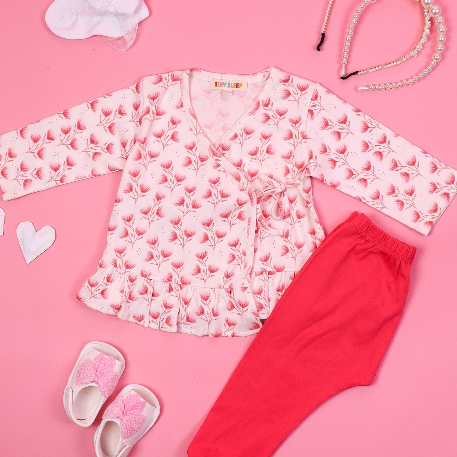 Baby girl's pink floral night suit set with a wrap-style top and matching red pants, displayed on a pink background with accessories like pearl necklaces, baby shoes, and a hairband.