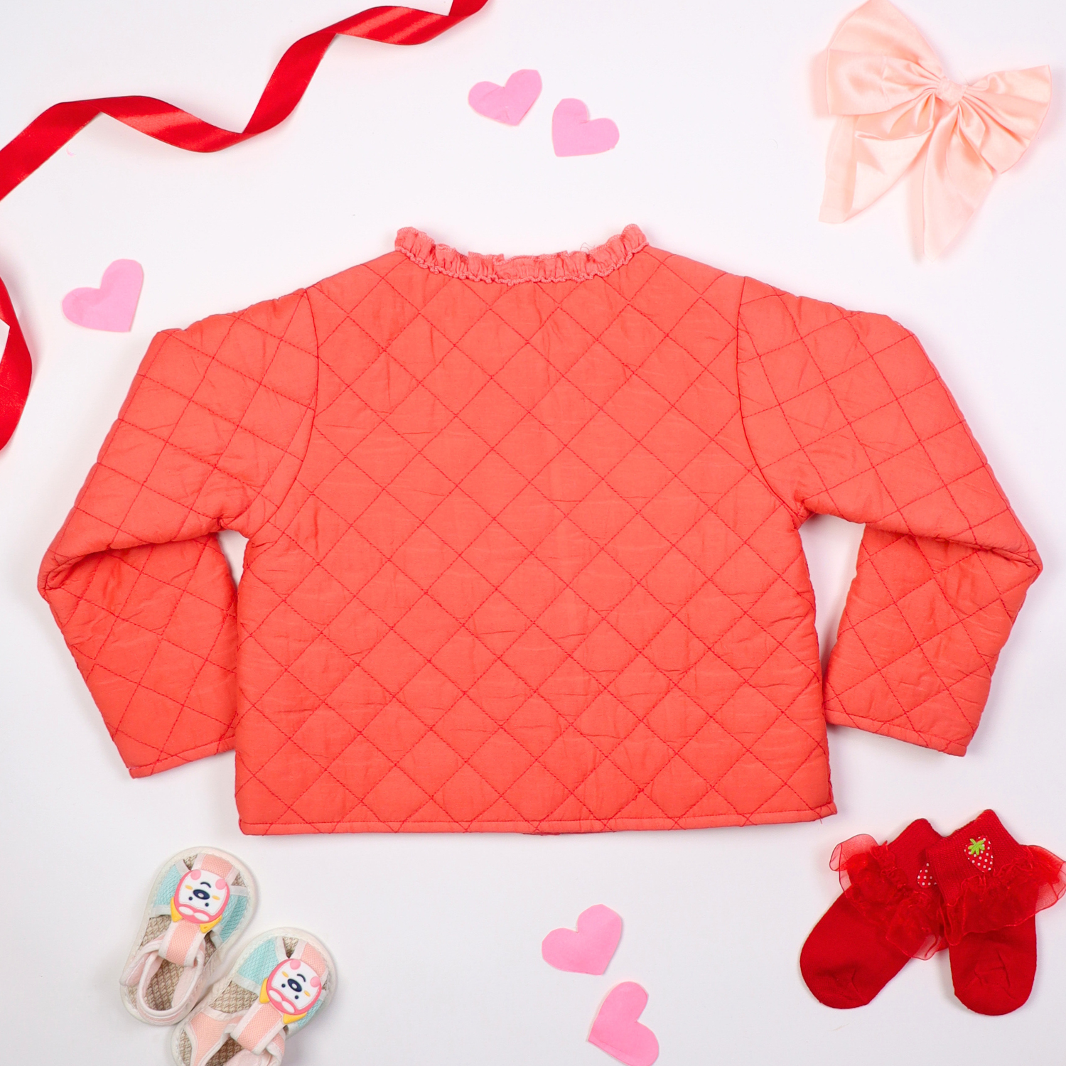 Back view of pink quilted baby jacket with ruffle details and heart-shaped pockets, displayed on a white background with decorative elements like ribbons, hearts, socks, and baby shoes.