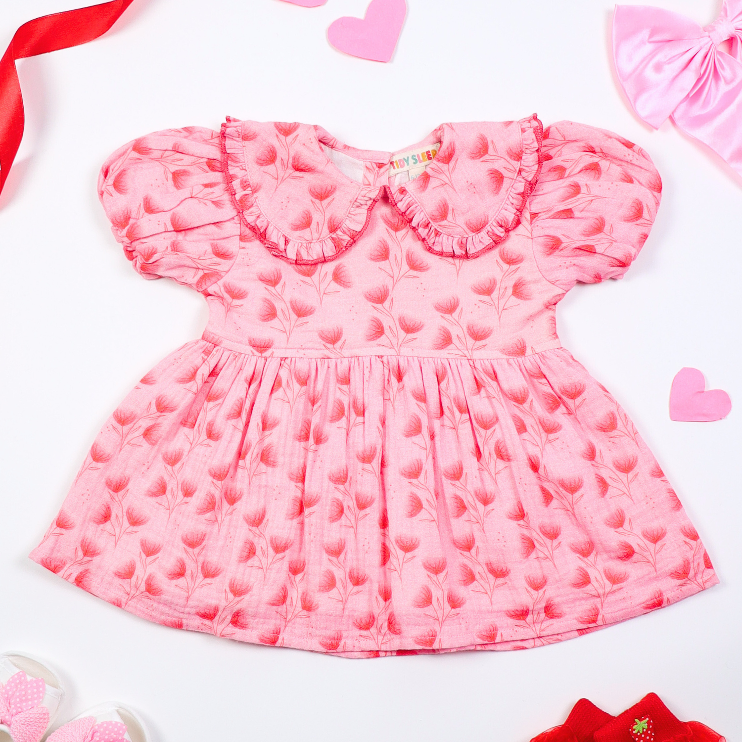 Pink floral baby dress with puff sleeves and a ruffled collar, displayed on a white background with decorative elements like ribbons, hearts, and baby shoes.