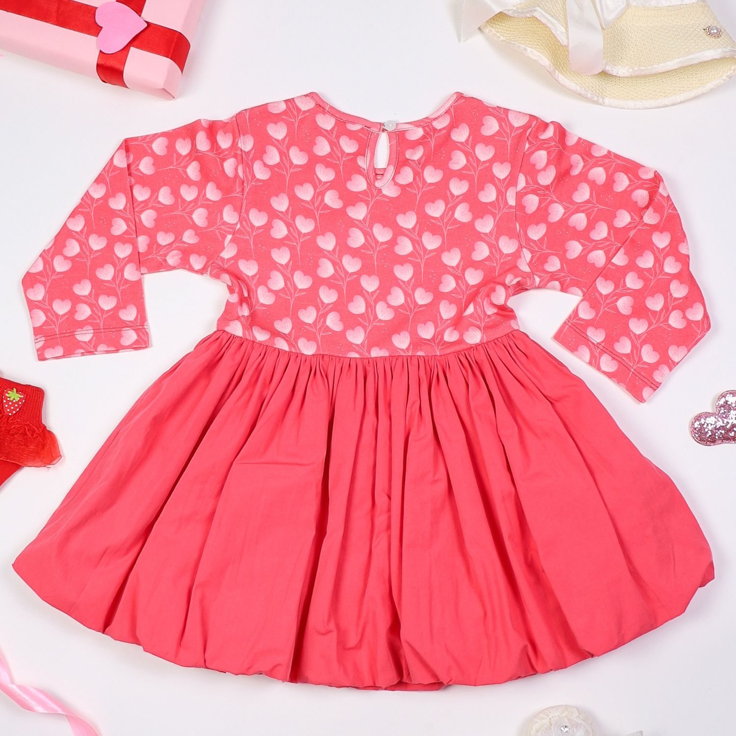 Back view of pink long-sleeve baby dress with a heart print top and flared skirt, displayed on a white background with decorative elements like ribbons, socks, and gift wrapping.