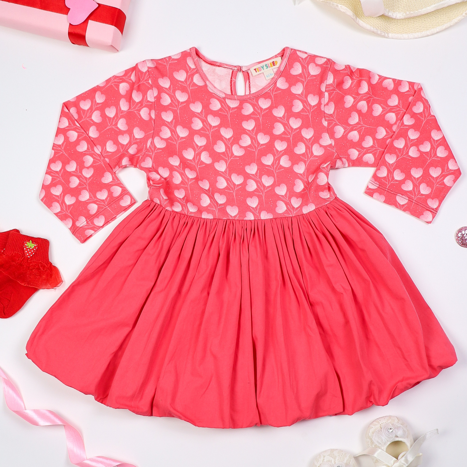 Pink long-sleeve baby dress with a heart print top and flared skirt, displayed on a white background with decorative elements like ribbons, socks, and gift wrapping.