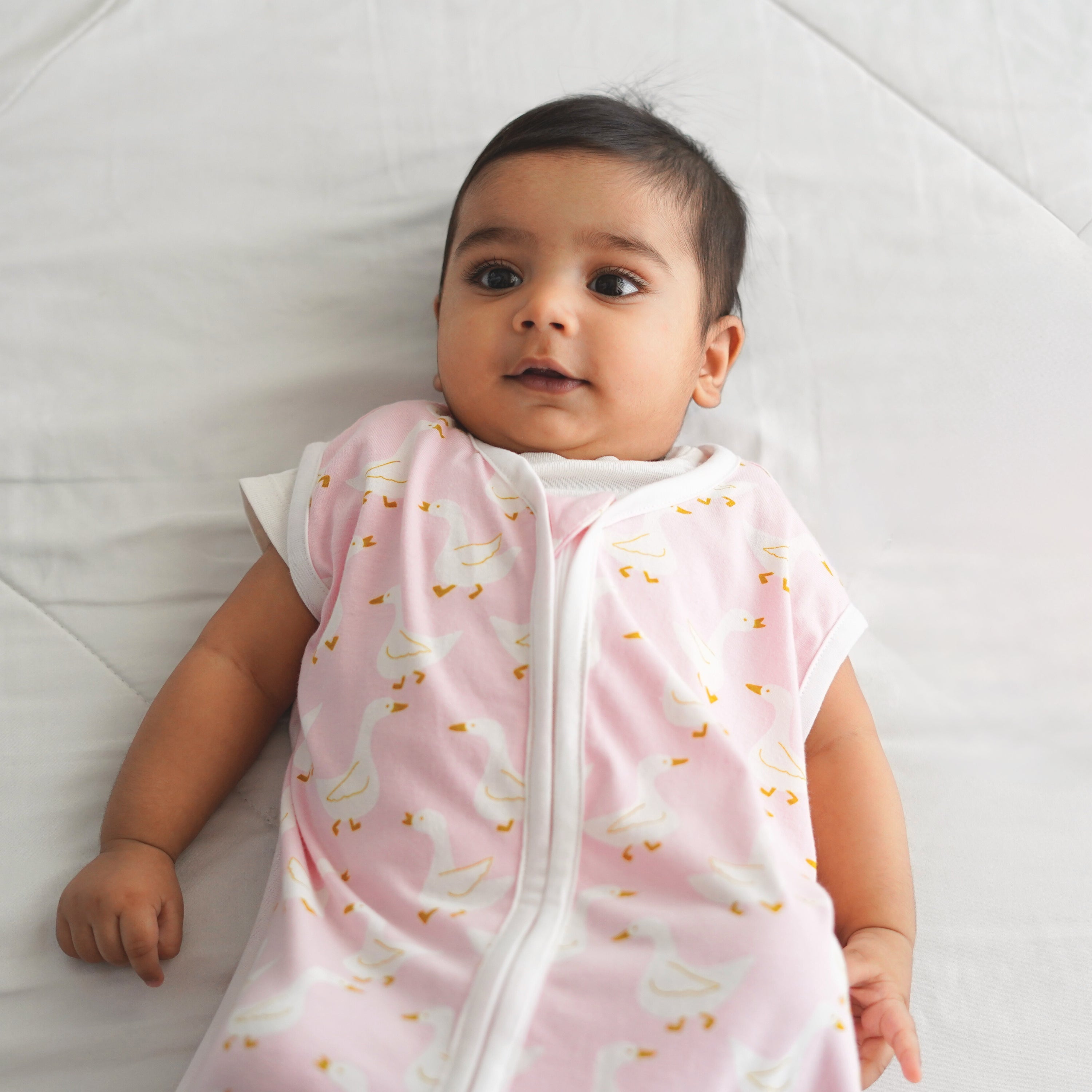 A baby lying on a white quilted surface, wearing a light pink sleeveless onesie with a white zipper and a playful goose pattern.