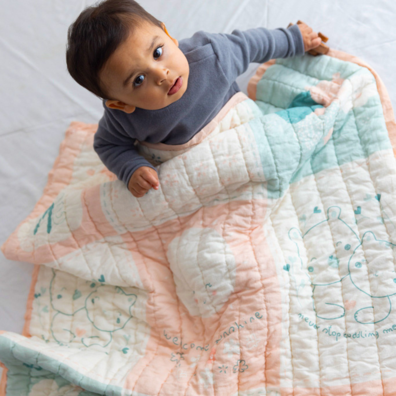 Baby wrapped in a soft pastel quilt with  bear illustrations and embroidered text, looking up with curiosity.