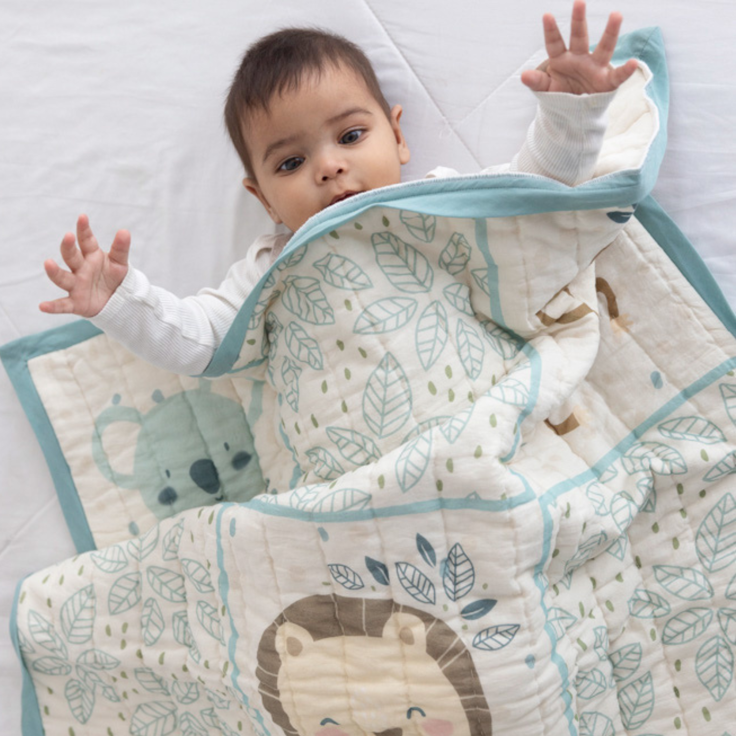 Baby lying on a bed, wrapped in a cozy quilt with adorable animal and leaf prints, raising hands playfully.